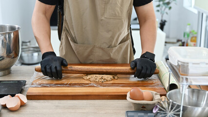 A man is making a dough on a wooden board. He is wearing black gloves. There are several bowls and cups on the table.