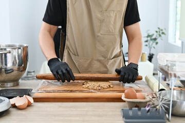 A man is making a dough on a wooden board. He is wearing black gloves. There are several bowls and cups on the table.