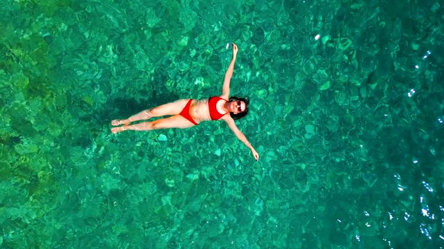Woman in red swimsuit lays on her back in the clear sea, top down view