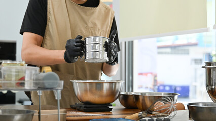A man is making a cake and is using a sifter to sift flour. The kitchen is well-equipped with various bowls, spoons, and other utensils. The atmosphere is calm and focused.