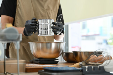 A man is making a cake and is using a sifter to sift flour. The kitchen is well-equipped with various bowls, spoons, and other utensils. The atmosphere is calm and focused.
