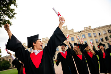 Excited young graduates celebrate success on a sunny campus day outdoors, proudly wearing caps and...