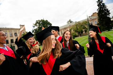 Excited graduates celebrate together in their black mantles on a sunny day, showcasing joy and...