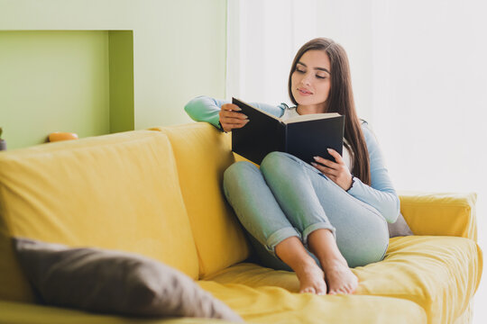 Young woman relaxing at home while reading a book on a cozy yellow sofa in a bright and comfortable living room setting.