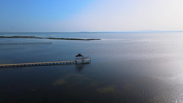 An aerial view of a serene dock extending into the calm waters of Coucou Beach in Bizerte. The wooden pier leads to a small hut, creating a peaceful and inviting scene for visitors.