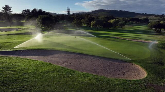 Low aerial orbits sprinklers watering golf green in golden hour light