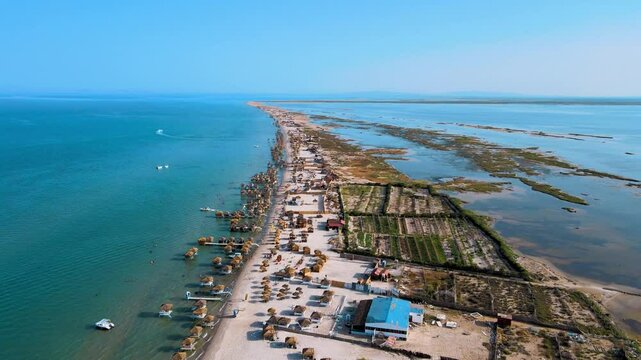 An aerial shot of Coucou Beach in Sidi Ali Makki, Bizerte. The long stretch of sandy beach is lined with numerous straw huts and boats, set against the backdrop of clear blue waters.