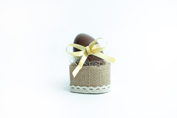 A chocolate Easter egg with a golden ribbon sits in a rustic burlap basket with lace detailing, creating a stylish and festive holiday presentation against a clean white background.
