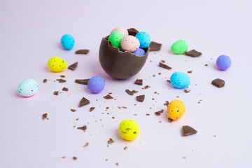 A cracked chocolate egg filled with colorful candy-coated mini eggs, surrounded by scattered chocolate pieces. A festive Easter concept on a pastel background.