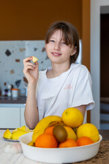 Obraz premium Smiling girl eating fresh fruit in kitchen. A cheerful young girl in a white shirt sits at a kitchen table, holding a piece of fruit while surrounded by a bowl of fresh fruits.