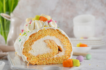 Easter carrot roll cake with cream cheese frosting, decorated chocolate eggs, jellied carrots and sugar sprinkles on top. Traditional carrot roll cake on a concrete background. Easter festive dessert.