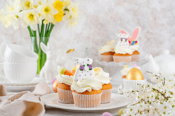 Easter cupcakes are decorated with cream, sugar bunnies, chocolate eggs, jelly carrots on a white plate. Easter food composition. Copy space.