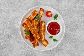 Fried halloumi cheese sticks with tomato sauce on a white plate on a concrete background.
