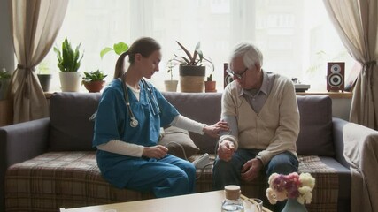Nurse in blue scrubs checking blood pressure of elderly man while sitting on couch in cozy living room, providing medical care and support in retirement home - Powered by Adobe
