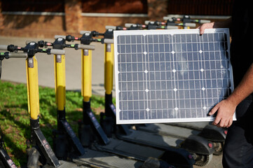 Male person holding solar panel in front of line of electric scooters parked in park. Integration of sustainable renewable energy, showcasing clean energy solution for charging electric scooters.