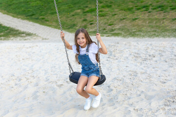Happy joyful little girl in white summer dress rides on a swing on a sandy beach with green lawn background or on playground in children park, on a sunny day. Vacation concept. Children's Day
