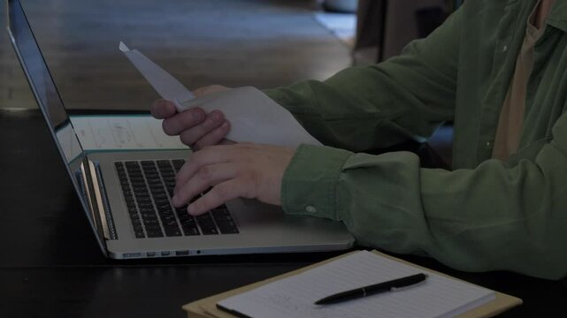 A middle-aged man in a green shirt calculating taxes and fees from a magazine with a laptop and writing notes at a table at home.