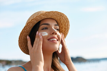 A young woman enjoys her day at the beach while applying sunscreen on her face, embracing sun...