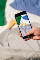Woman hand with smartphone taking picture of used shirt. Preparing clothes to sell in second hand online shop. Resell goods and Second hand concept. Clothes donation
