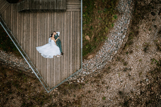 Aerial view of a newlywed couple lying on a wooden deck, embracing and enjoying a romantic moment in a natural, scenic outdoor setting..