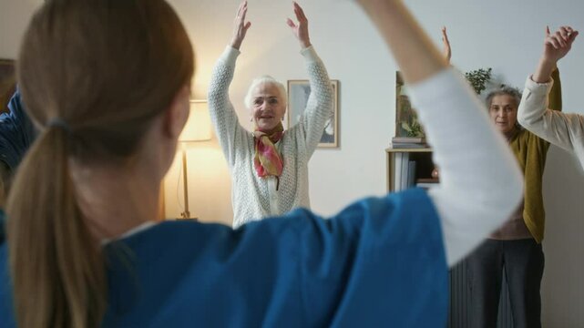 Diverse elderly people following caregiver doing light exercise during group fitness session in living room of nursing home