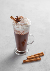 Hot chocolate with whipped cream, cinnamon and spices in a tall glass on a light background.