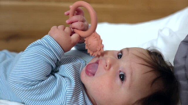 Baby lying on soft bed chewing teething ring with hedgehog design, gripping toy with both hands, soothing gums, exploring textures, engaging in oral sensory development