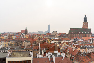 Obraz premium Panoramic view of historic European city with red rooftops, Gothic church, and modern skyscraper in the background on cloudy day. Architectural contrast between old and new urban landscapes.