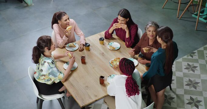 Indian young adult woman group old lady sitting around table talk gossip eat break fast food share lunch at job work place happy female girl drink juice pick up coffee cup have fun joy spend day time