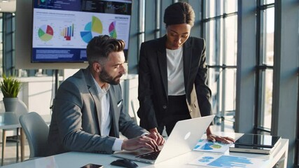 Two professionals analyzing business charts and graphs on laptop and large screen display, with a man seated and a woman standing, working together in a bright, modern office with large windows