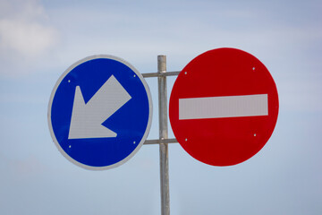 Two circular road signs are prominently displayed against a pale blue sky. On the left, a blue sign features a white arrow pointing downwards and to the left, indicating a direction.