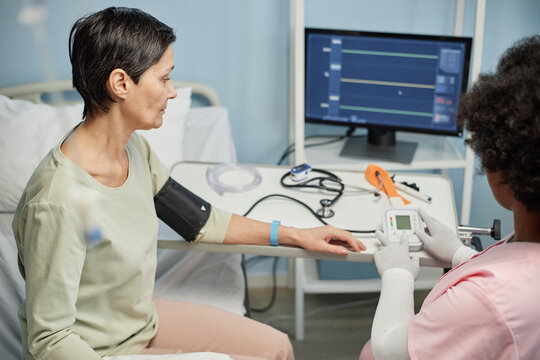 Healthcare worker caring for patient by measuring blood pressure with medical equipment in a hospital room Patient seated on hospital bed during examination, suggesting routine health checkup