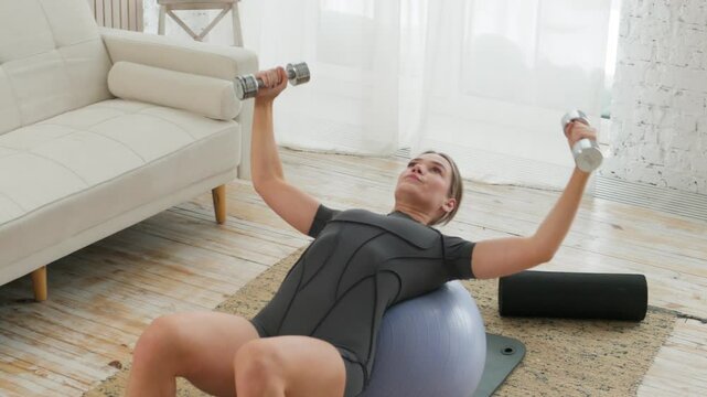 Young caucasian woman doing back strengthening and biceps exercises lying on her back on a gym ball and holding dumbbells in her hands in a bright room. The concept of a healthy lifestyle. Sports.