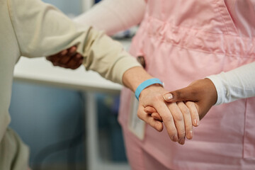 Close-up of a caregiver in pink uniform gently holding hands with an elderly person during a comforting interaction in a healthcare setting