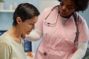 Compassionate nurse dressed in pink scrubs assisting elderly patient during medical checkup in clinic room with medical equipment visible in background