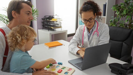 Doctor consulting father with young son in a clinic office, using a laptop while discussing patient care during a medical appointment, highlighting family interaction indoors.