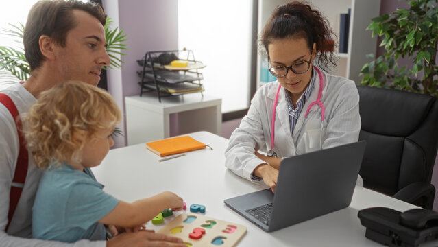 Doctor consulting father with young son in a clinic office, using a laptop while discussing patient care during a medical appointment, highlighting family interaction indoors.