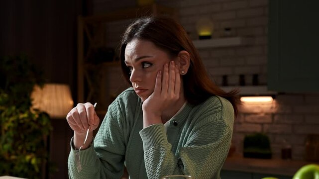 Woman at the table in the kitchen preparing healthy vegetable salad for dinner, woman mixing salad with fork on the plate, no appetite, sad unhappy expression. Slow motion
