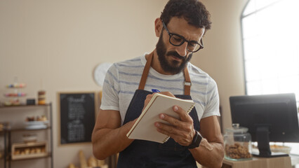 Young hispanic man holding a notebook writes in a bakery interior with shelves of pastries in the...