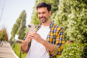 Young man enjoying outdoors using smartphone, dressed in casual checkered shirt, smiling in...