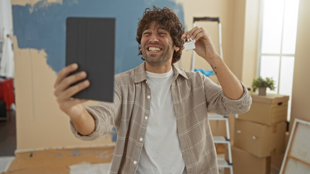 Young man smiling indoors holding keys and taking a photo in his new home with unfinished walls and moving boxes around, capturing a joyful moment of homeownership.