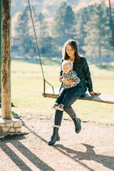 Little girl sitting on her mother lap swinging on a wooden swing in the park