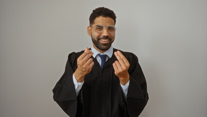 Young, hispanic, man in a graduation gown making a money gesture with his fingers, isolated over a white background, smiling and looking directly at the camera