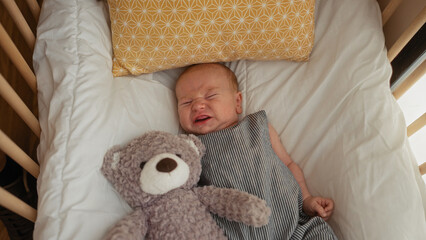 Newborn baby boy crying in a crib next to a teddy bear in a cozy bedroom setting