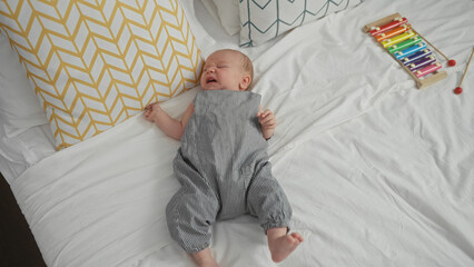 Naklejka premium Crying baby boy lying on a bed in a bedroom dressed in striped romper with pillows and a xylophone nearby