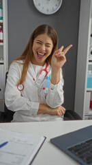 Woman doctor in clinic room smiling and showing victory gesture while seated at desk wearing white...