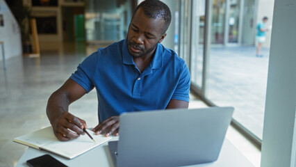 Young man working in office with laptop writing in notebook while wearing a blue polo