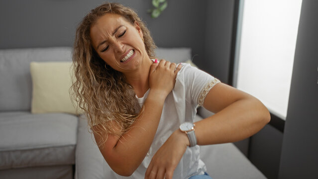 Woman experiencing shoulder pain sitting in a living room, expressing discomfort in a home interior setting with a grey couch in the background.