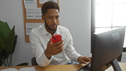Young man working in an office, looking at his smartphone while using a computer, wearing a white shirt, indoor setting, documents pinned on noticeboard, plants in the background, modern workspace