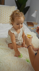 Fototapeta premium Cute blonde toddler girl sitting on a bed in a cozy bedroom, being fed with a spoon by an adult, capturing a moment of childhood in an indoor home setting.